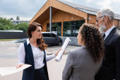 real estate broker with papers talking to interracial business partners near blurred building outdoors