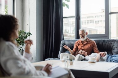 Orta yaşlı bir adam bulanık Afrikalı Amerikalı psikologla konuşuyor. Kanepede gülümseyen karısının yanında.