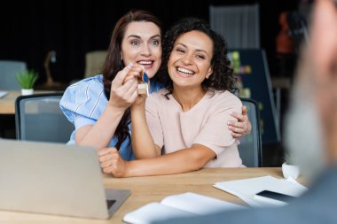 happy multiethnic lesbian couple holding key near blurred broker in real estate agency clipart