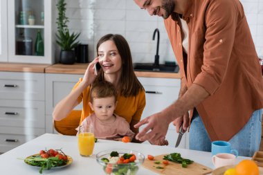 Akıllı telefondan konuşan genç bir kadın oğlu ve kocası evde salata pişiriyor. 