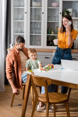 Akıllı telefondan konuşan, dizüstü bilgisayarın yanında bardak tutan ve evde oyun oynayan bir kadın. 