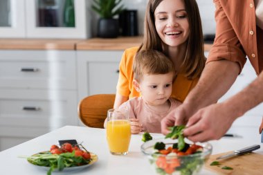 Mutlu kadın oğlunun yanında oturuyor ve kocası mutfakta salata pişiriyor. 