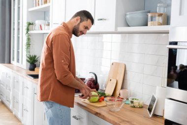 Mutfakta sebze keserken dijital tablete bakan gülümseyen adamın yan görüntüsü 