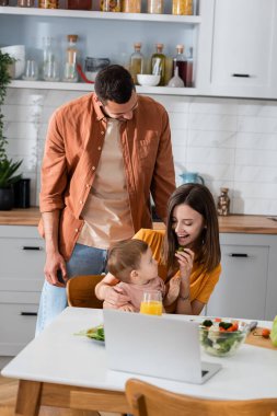 Pozitif aile, dizüstü bilgisayarın yanında küçük bir çocuğa bakıyor ve mutfakta salata var. 
