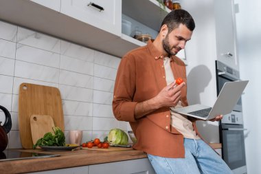 Mutfakta sebzelerin yanında kirazlı domates ve laptop tutan genç adam. 