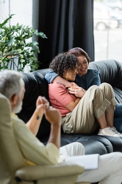 Glada Mångkulturella Lesbiska Par Omfamna Soffan Nära Suddig Psykolog — Stockfoto