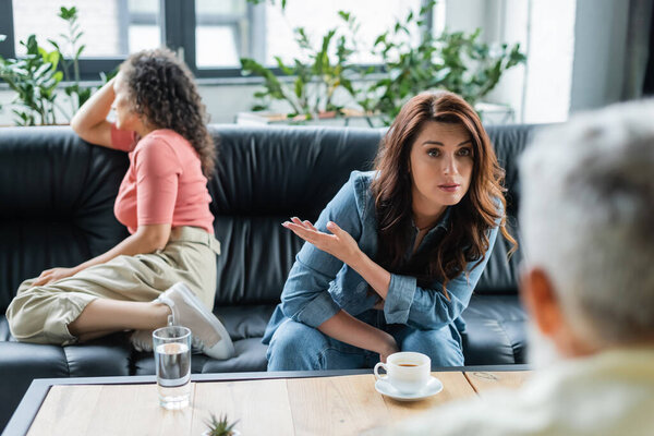 lesbian woman pointing at depressed african american girlfriend while talking to blurred psychologist
