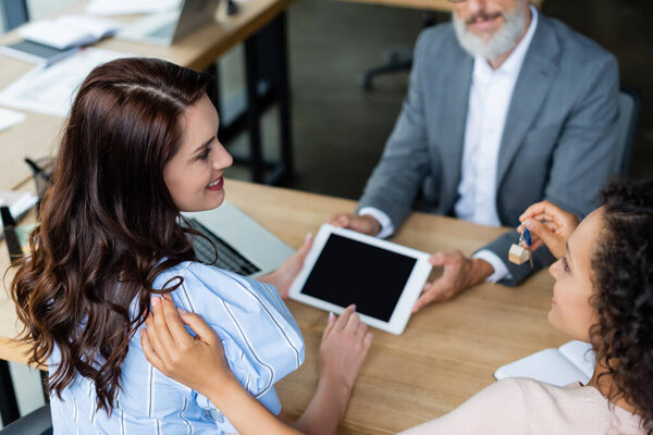 african american woman holding key near happy lesbian girlfriend and blurred realtor with digital tablet