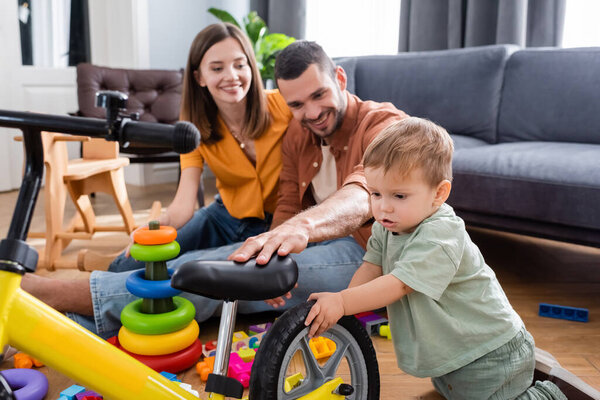 Kid standing near bike and smiling parents in living room 