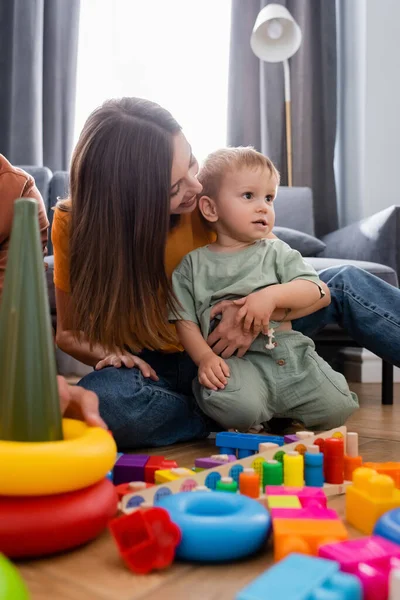 Sonriente Mujer Abrazando Hijo Cerca Borrosa Juguetes Sala Estar —  Fotos de Stock