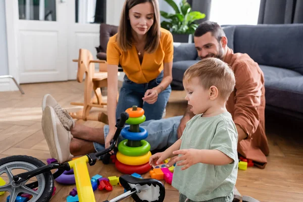 Toddler kid standing near bike, toys and blurred parents in living room