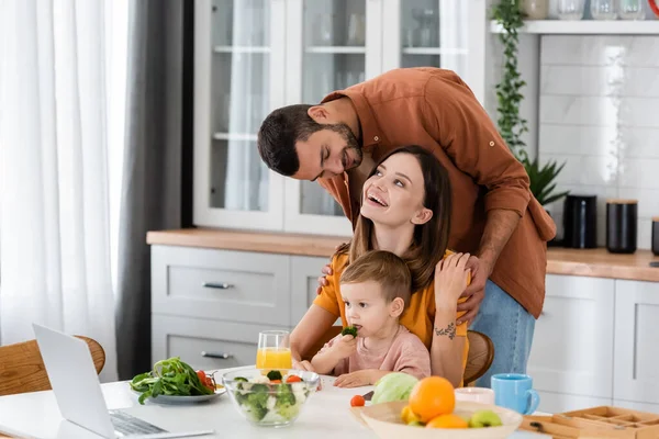 Hombre Positivo Abrazando Esposa Cerca Hijo Ensalada Portátil Cocina — Foto de Stock