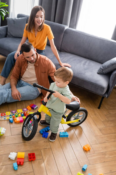 Toddler boy standing near bike and parents in living room 