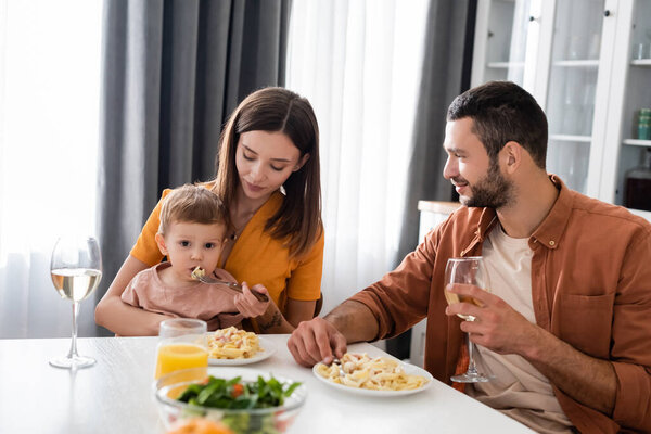 Man holding glass of wine while wife feeding son with pasta at home 