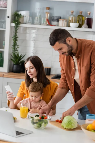 Hombre Cocina Ensalada Cerca Esposa Usando Teléfono Inteligente Hijo Pequeño — Foto de Stock