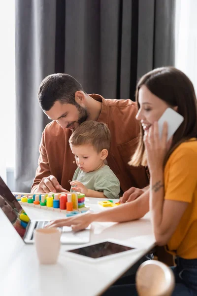 笑顔の男は息子の近くに座って教育ゲームをプレイし 妻はノートパソコンで作業し 自宅で携帯電話で話す — ストック写真