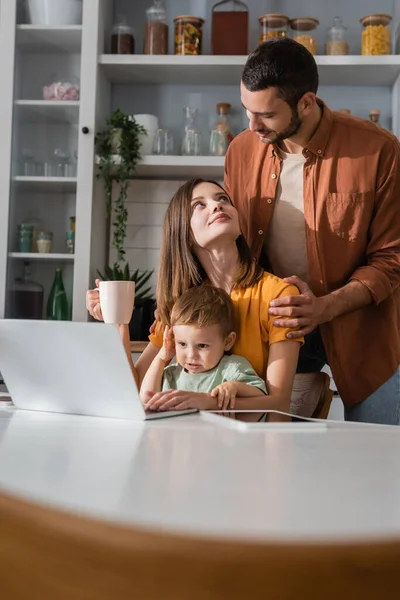 Joven Abrazando Esposa Con Taza Cerca Hijo Gadgets Casa —  Fotos de Stock