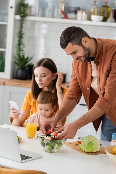 Man Matlagning Sallad Nära Laptop Och Suddig Familj Köket — Stockfoto