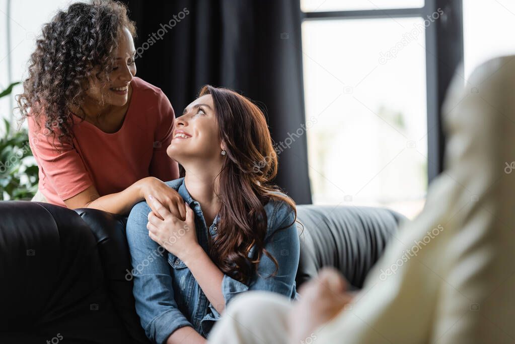 happy same sex couple looking at each other during consultation with psychologist on blurred foreground