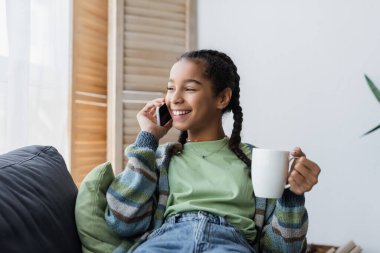 Neşeli Afrikalı Amerikalı genç kız elinde çay bardağıyla evde akıllı telefondan konuşuyor.