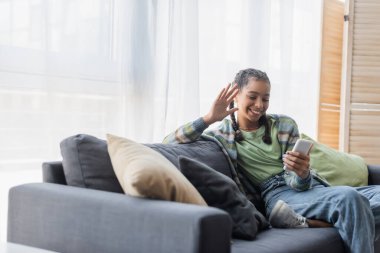 Neşeli Afro-Amerikalı genç kız akıllı telefondaki video görüşmesi sırasında kanepede oturup el sallıyor.