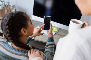 Afrikalı Amerikalı bir kız bilgisayar ekranının yanında boş ekranlı cep telefonu kullanıyor ve annesini bir fincan çayla bulanıklaştırıyor.