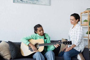 Gülümseyen Afro-Amerikan kadın. Kanepede oturan genç kız gitar çalıyor.