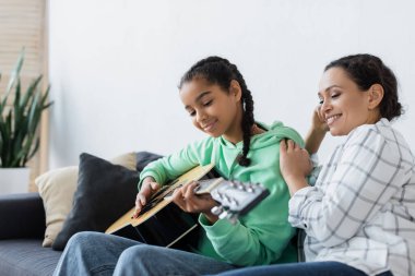 Afro-Amerikalı genç kız evde gülümseyerek gitar çalıyor.