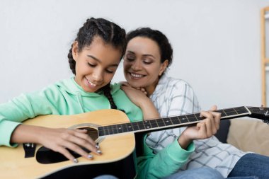 Mutlu bir Afro-Amerikan kadını genç kızının yanında gülümsüyor ve evde gitar çalıyor.