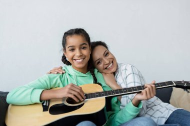 Mutlu Afro-Amerikan kadını gülümseyen kızını kucaklıyor ve evde gitar çalıyor.
