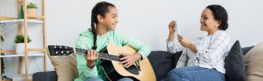 Cep telefonlu, gülümseyen kızının evde gitar çalarken fotoğrafını çeken Afro-Amerikan bir kadın.