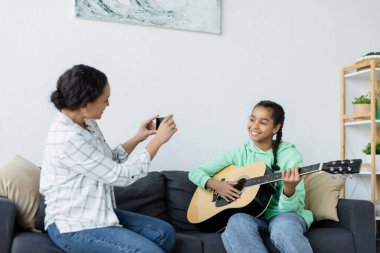 Neşeli Afrikalı Amerikalı genç kız annesinin yanında gitar çalıyor cep telefonuyla fotoğraf çekiyor.