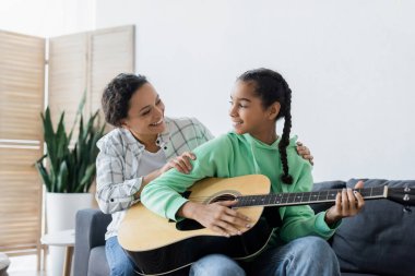 Gülümseyen Afrikalı Amerikalı kız evde gitar çalarken mutlu anneye bakıyor.