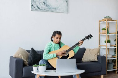 Genç Afrikalı Amerikalı kız yastıklı koltukta otururken dizüstü bilgisayarın yanında gitar çalmayı öğreniyor. 