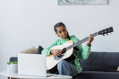 Afro-Amerikan genç kız dizüstü bilgisayarın yanında oturuyor ve gitar çalmayı öğreniyor.