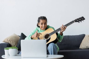 Gülümseyen Afrikalı Amerikalı kız dizüstü bilgisayara bakıyor ve evde çalmayı öğrenirken gitara işaret ediyor.