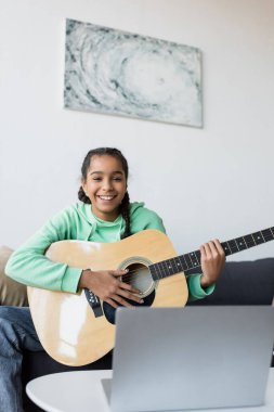 Akustik gitarlı neşeli Afro-Amerikalı genç kız bulanık dizüstü bilgisayarın yanındaki kameraya bakıyor.