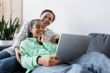 Genç kızı olan neşeli Afrikalı Amerikalı kadın bulanık laptoptan film izliyor.