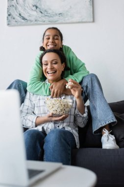 Gülümseyen Afro-Amerikan kızı, elinde patlamış mısırla gülen annesini kucaklarken bulanık dizüstü bilgisayardan film izliyor.