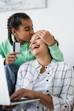 Afro-Amerikalı bir kız. Annesinin mutlu alnını öpüyor. Kredi kartını bulanık dizüstü bilgisayarın yanında tutuyor.