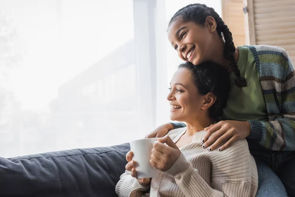 快乐的非洲裔美国少女抱着快乐的母亲坐在沙发上 手里拿着茶杯 — 图库照片