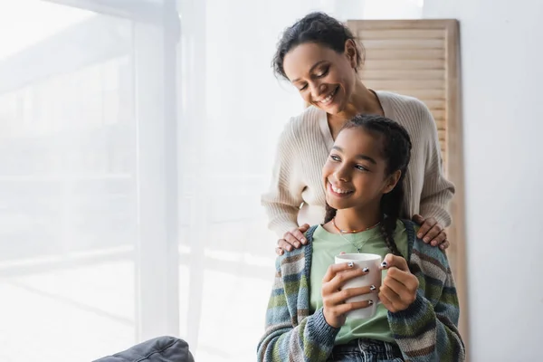 Pleased African American Woman Hugging Shoulders Teenage Daughter Holding Tea — Stock Photo, Image