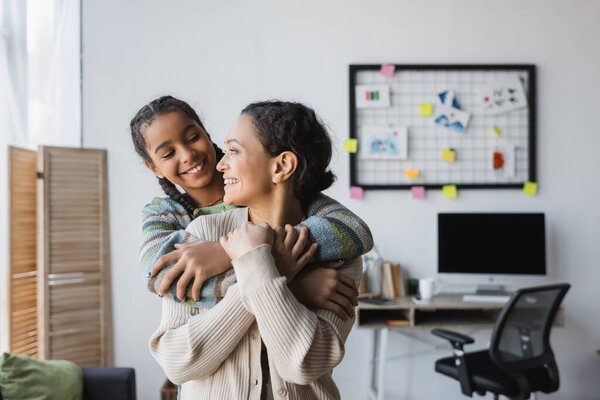 cheerful african american mother and daughter looking at each other while embracing at home