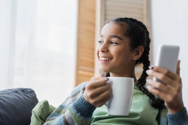 Adolescente Afroamericana Complacida Sosteniendo Una Taza Teléfono Inteligente Borroso Casa — Foto de Stock