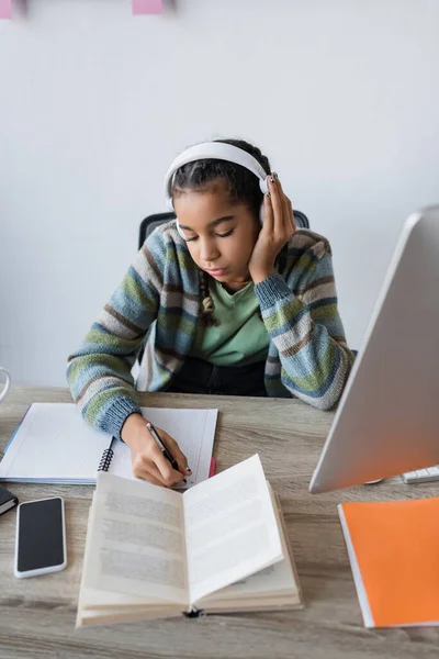 戴耳机的非洲裔美国女孩 在电脑旁边的笔记本电脑上写作 而智能手机旁边的屏幕则是空白的 — 图库照片