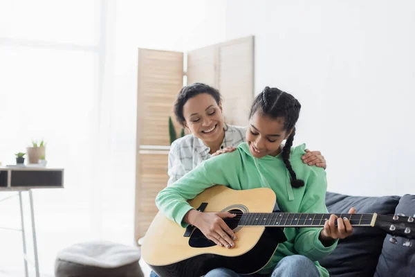 Africano Americano Chica Jugando Guitarra Mientras Sentado Sofá Cerca Mamá —  Fotos de Stock