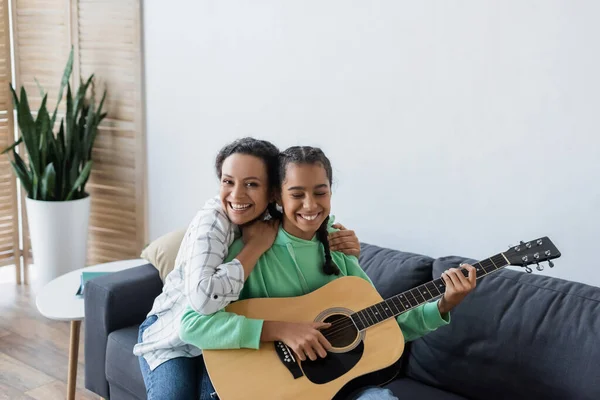 Alegre Afroamericana Mujer Mirando Cámara Mientras Abrazando Hija Jugando Guitarra —  Fotos de Stock