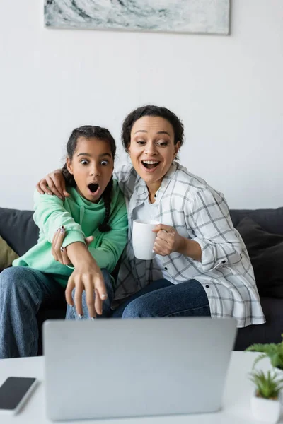 震惊的非洲裔美国女孩一边和兴奋的妈妈一起看电影 一边指着模糊的笔记本电脑 — 图库照片