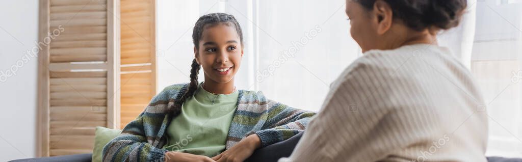 smiling african american girl looking at blurred mother during conversation at home, banner