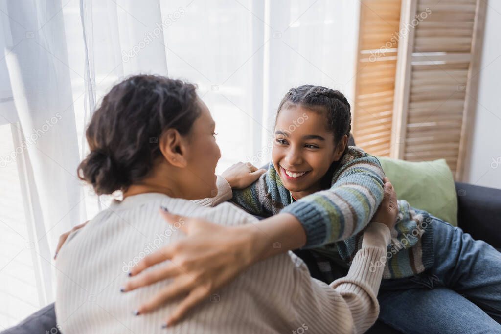 pleased african american teenage girl hugging blurred mom on couch at home